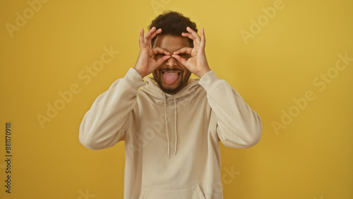 Playful african man making glasses with fingers, sticking tongue out against yellow background.