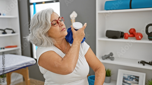 A mature woman applies ice to her shoulder in a therapeutic clinic's rehabilitation room.