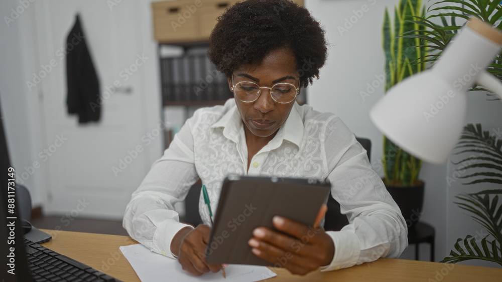 African american woman with curly hair working in an office, reading a tablet, and taking notes on paper in a modern indoor workspace. African american woman with curly hair working in an office, reading a tablet, and taking notes on paper in a modern indoor workspace.