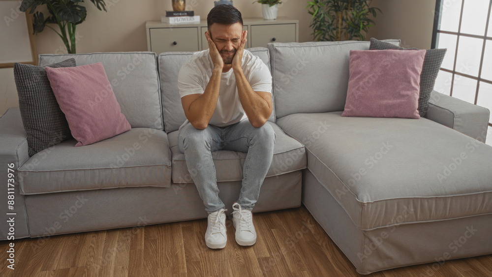 Young hispanic man with a beard sits on a grey couch in a modern living room, indoors, looking distressed and holding his face in his hands. Young hispanic man with a beard sits on a grey couch in a modern living room, indoors, looking distressed and holding his face in his hands.