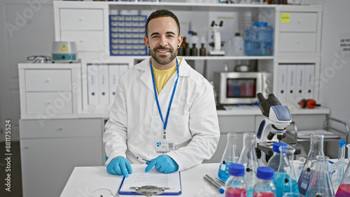 Handsome young man in labcoat writing notes in a clinical laboratory setting with microscope and vials present.