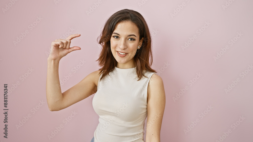 A smiling young hispanic woman gestures 'small' with her fingers against a pastel pink background, exuding beauty and positivity. A smiling young hispanic woman gestures 'small' with her fingers against a pastel pink background, exuding beauty and positivity.