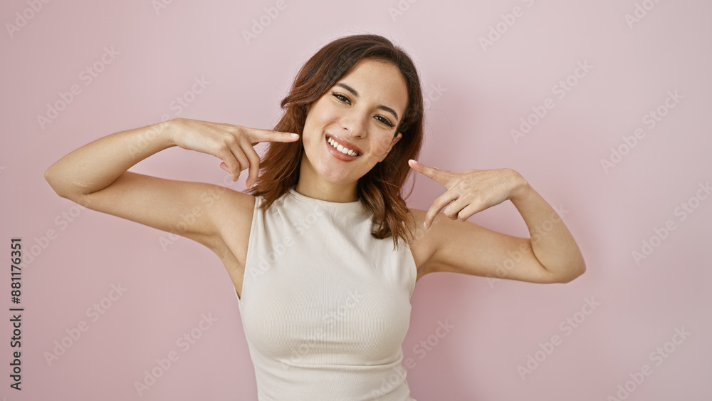 A smiling young hispanic woman pointing to her smile, isolated on a pink background, with a stylish hairstyle. A smiling young hispanic woman pointing to her smile, isolated on a pink background, with a stylish hairstyle.