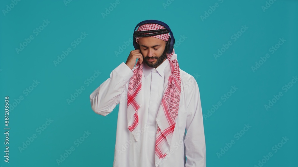 Fototapeta premium Joyful person dancing on songs in studio, listening to modern music on headphones and having fun on camera. Middle eastern adult in traditional clothing showing groovy dance moves.