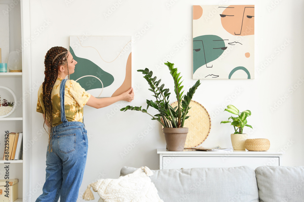 Young woman with dreadlocks hanging painting on wall at home Young woman with dreadlocks hanging painting on wall at home