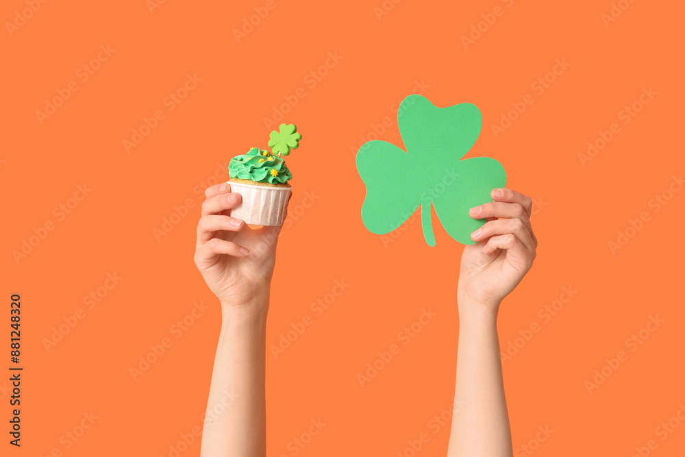 Female hands with tasty cupcake and paper clover for St. Patrick's Day on orange background Female hands with tasty cupcake and paper clover for St. Patrick's Day on orange background