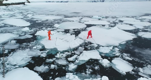 People in orange immersion suits run melting ice, Antarctica. Men in wetsuits try to escape from polar ocean trap, fall in cold water ice floes. Explore climate, global warming, melt icebergs. Aerial