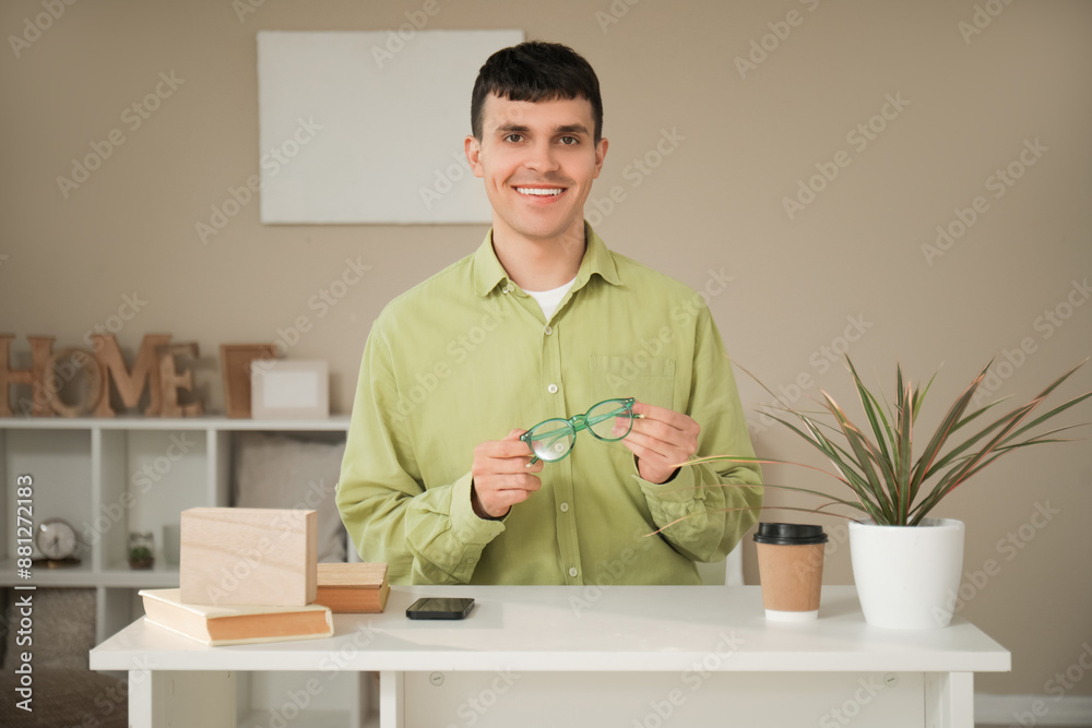 Handsome happy young man with new stylish eyeglasses sitting at table in room
