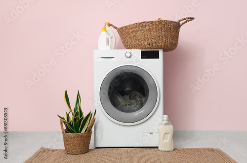 Modern washing machine with laundry, basket and houseplant near pink wall