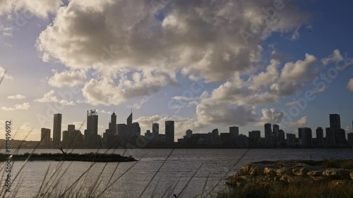 Perth City at dusk beneath swirling cumulous grey clouds - timelapse of view through swaying reeds past small island across Swan River. Black swan swims past