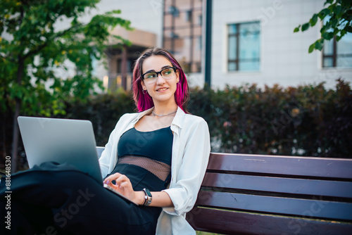 Portrait of a young beautiful female student sitting with a laptop on a bench outdoors. Long-haired brunette working remotely with a wireless portable computer. Mobile office, freelance concept.