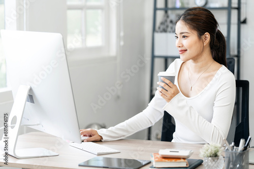 Beautiful young woman in casual clothing using desktop computer and enjoying hot drink while working indoors