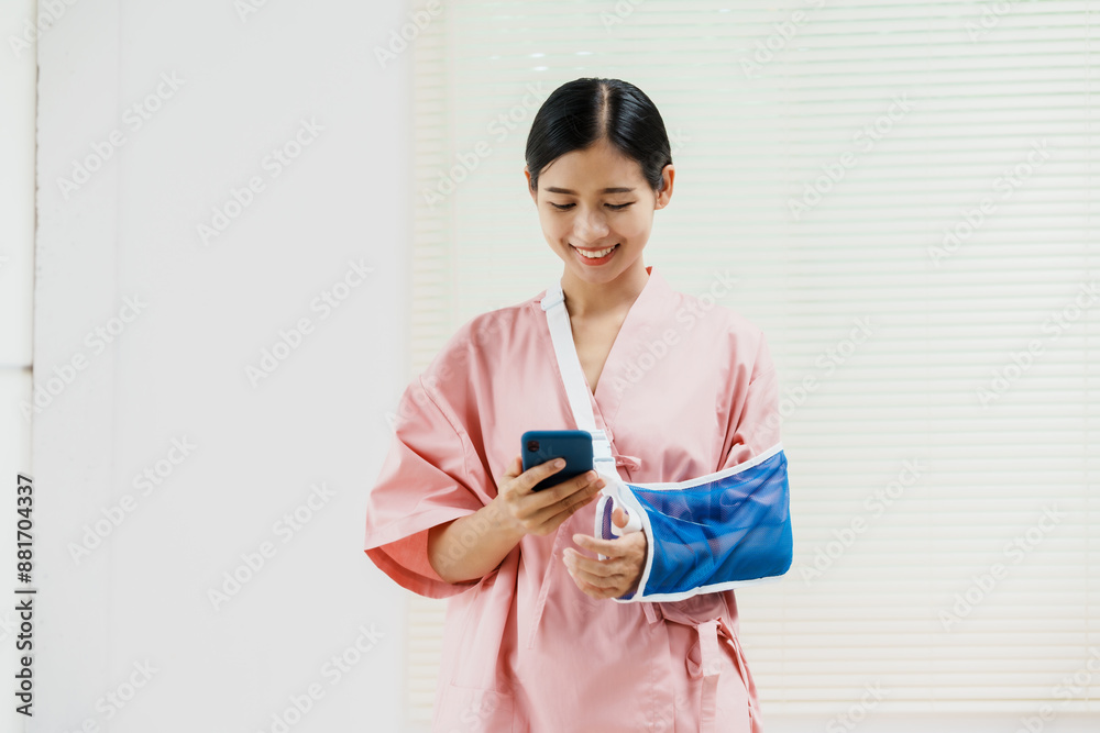 A young Asian female wearing a pink patient uniform stands isolated against a white wall background, showing confidence despite having her arm in a sling due to an injury. A young Asian female wearing a pink patient uniform stands isolated against a white wall background, showing confidence despite having her arm in a sling due to an injury.