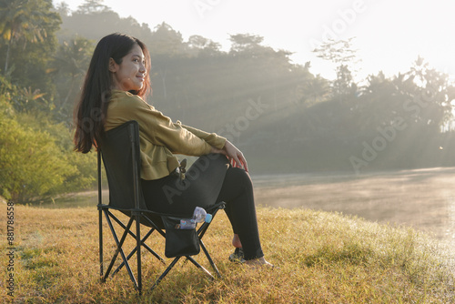 Peaceful Asian woman sits in a lawn chair by a lake, looking out at the water. The scene is serene with the sun shining through the trees and casting a warm glow on the water. photo