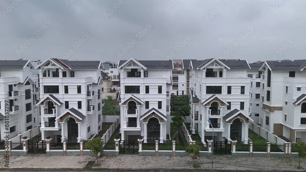 A newly constructed row of identical large white multi story villa styled houses along a suburban road. Drone flies up revealing rooftop view. A newly constructed row of identical large white multi story villa styled houses along a suburban road. Drone flies up revealing rooftop view.