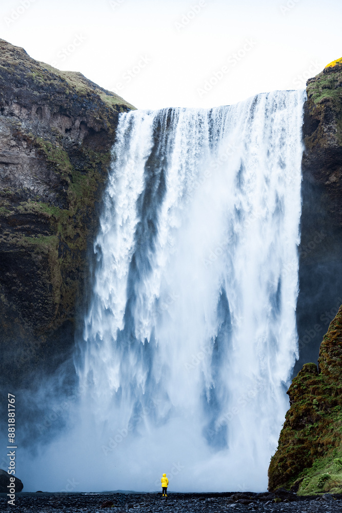 Stunning view of the Skógafoss Waterfall with a tourist wearing a yellow jacket. Skógafoss is one of the biggest waterfalls in Iceland, situated on the Skoga River in the south of Iceland.