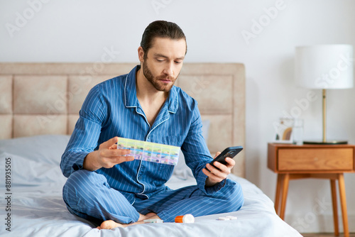 Handsome man in home seated on bed with attention fixed on cell phone and pill case. photo