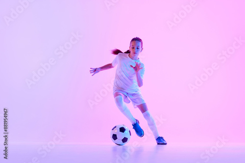 Concentrated teen girl, soccer player training, dribbling ball against pink studio background in neon light. Concept of sport, active and healthy lifestyle, childhood, school, hobby