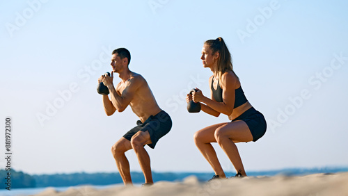 Canvas Print Athletic young man and woman with sportive body, in black sportswear doing squats with kettlebell, intensive outdoor training on beach by the river