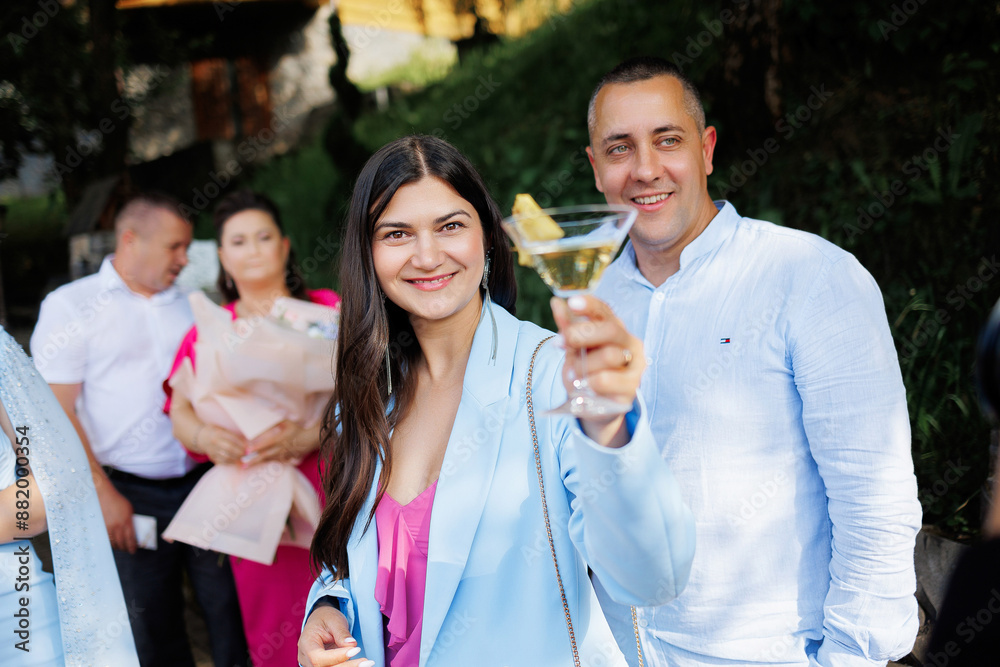 A man and woman are smiling and holding glasses of wine. The man is wearing a blue shirt and the woman is wearing a blue jacket. They are surrounded by other people, some of whom are holding handbags A man and woman are smiling and holding glasses of wine. The man is wearing a blue shirt and the woman is wearing a blue jacket. They are surrounded by other people, some of whom are holding handbags