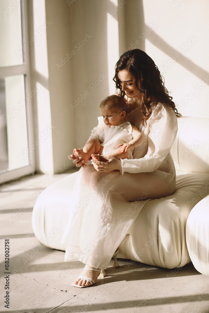 Happy mother in stylish dress sitting on the sofa and holding her baby daughter in her arms. Mother and daughter in same designed white dresses Happy mother in stylish dress sitting on the sofa and holding her baby daughter in her arms. Mother and daughter in same designed white dresses