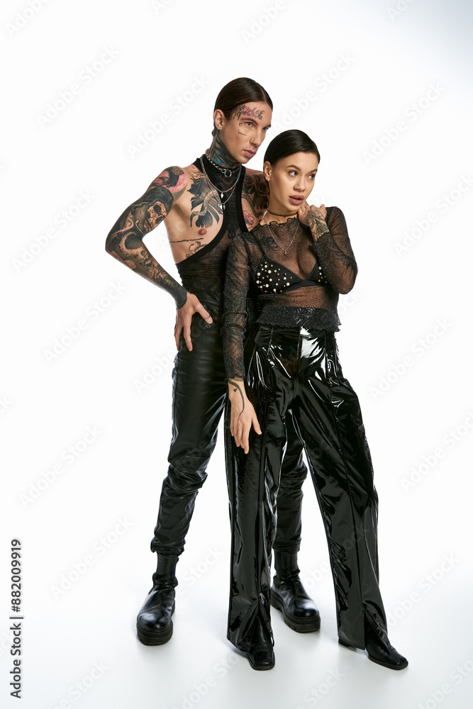 A young stylish couple with tattoos standing close together in a studio on a grey background. A young stylish couple with tattoos standing close together in a studio on a grey background.