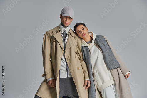 Fotografie A young, stylish couple stands confidently side by side in trench coats against a grey backdrop