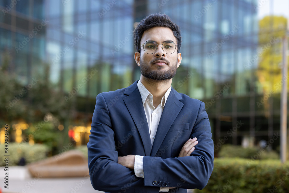 Confident businessman wearing suit standing with arms crossed outside modern office building. Professional appearance with eyeglasses, beard, and serious expression. Confident businessman wearing suit standing with arms crossed outside modern office building. Professional appearance with eyeglasses, beard, and serious expression.