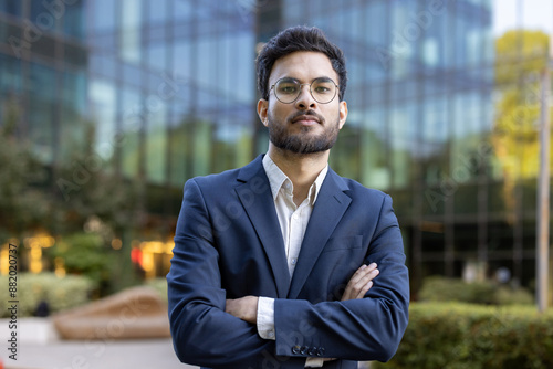 Wallpaper Mural Confident businessman wearing suit standing with arms crossed outside modern office building. Professional appearance with eyeglasses, beard, and serious expression. Torontodigital.ca
