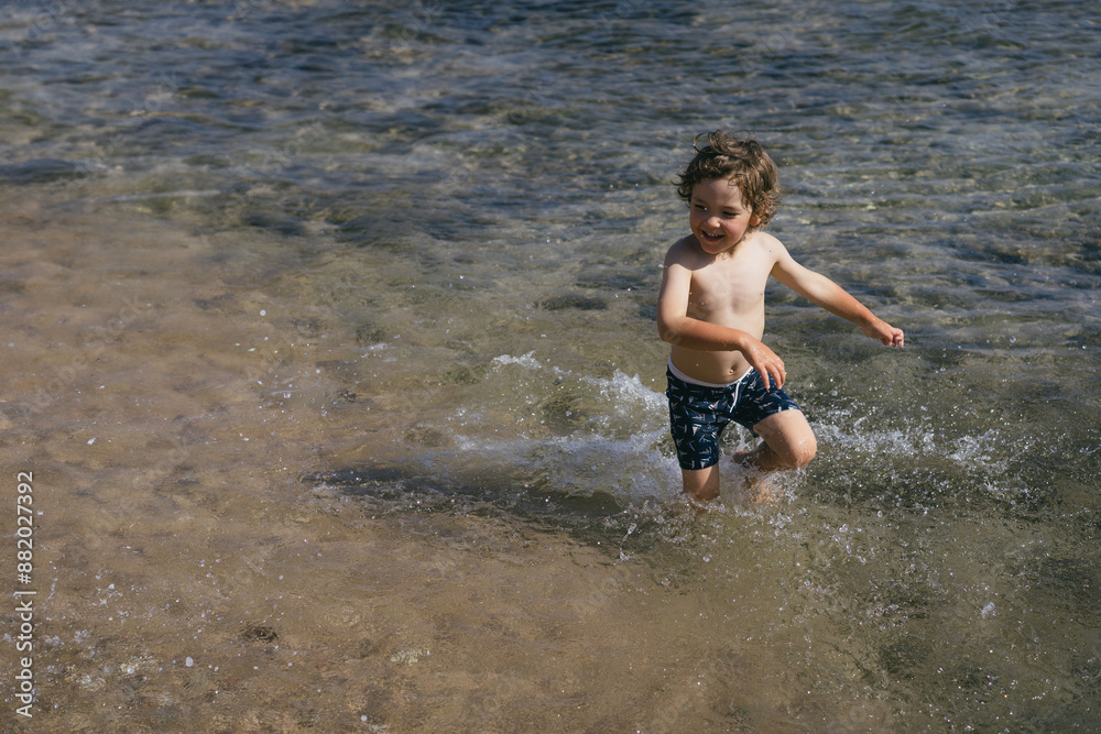 Fototapeta premium A young boy runs through the shallow water of the ocean on a sunny day. He is wearing blue swim trunks and is splashing water as he runs.