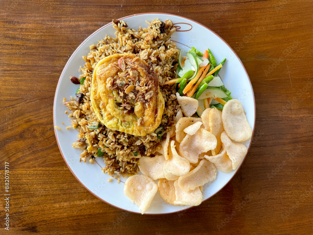 Local Indonesian ketchup fried rice with scrambled egg and prawn crackers, cucumber slices, served on top of white plate, on a wooden table. Local Indonesian ketchup fried rice with scrambled egg and prawn crackers, cucumber slices, served on top of white plate, on a wooden table.
