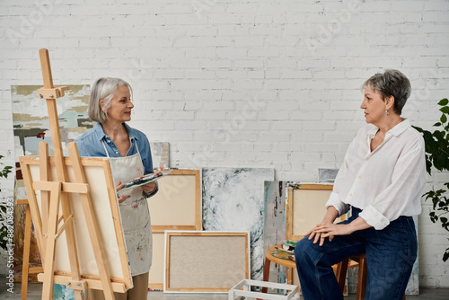 Two mature women in an art studio, one holds a paint palette while the other sits and observes.