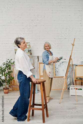 Two women in an art studio. One is painting on a canvas while the other leans against a stool, watching. © Bliss