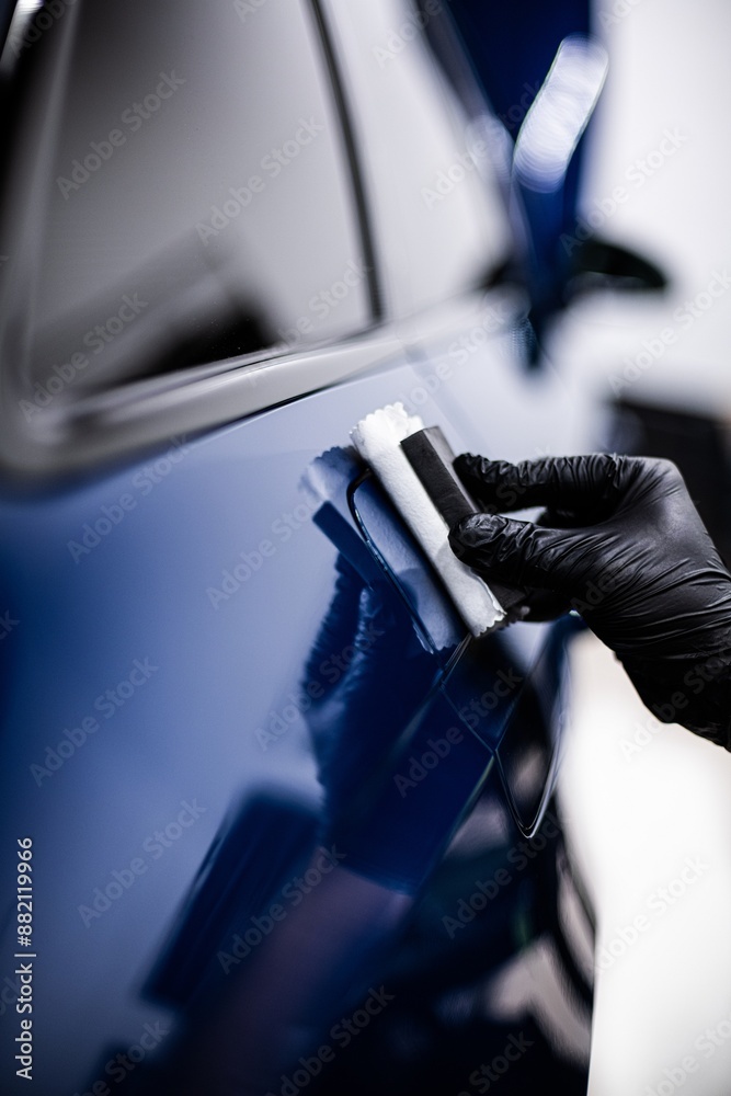 Image shows a person applying a ceramic coating to the surface of a blue car. The person is wearing black gloves, and the setting appears to be a well-lit workshop or garage