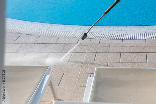 A high-pressure water jet cleaning the poolside area, enhancing the shine of the lounge chairs beside the vibrant blue water of the swimming pool.