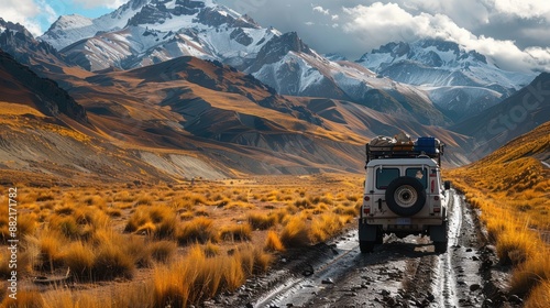 A rugged off-road vehicle traversing a muddy dirt path through a picturesque mountainous landscape with snow-capped peaks and dry grassland