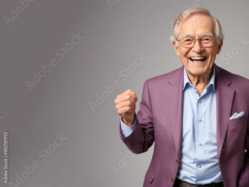 Older white senior man in suit smiles broadly, raising his fist in celebration against light gray background