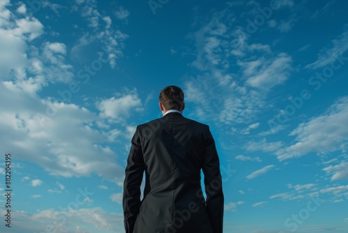 Back view of a businessman in a suit staring at a blue sky filled with white clouds, symbolizing prospects.