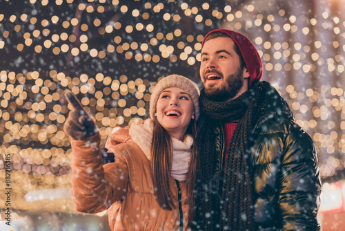 Photo of amazed girfriend point index finger evening illumination sky boyfriend look x-mas christmas magic atmosphere wear coat scarf hat