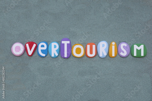 Overtourism, the congestion or overcrowding from an excess of tourists, resulting in conflicts with locals, creative text composed with multi colored stone letters over green sand