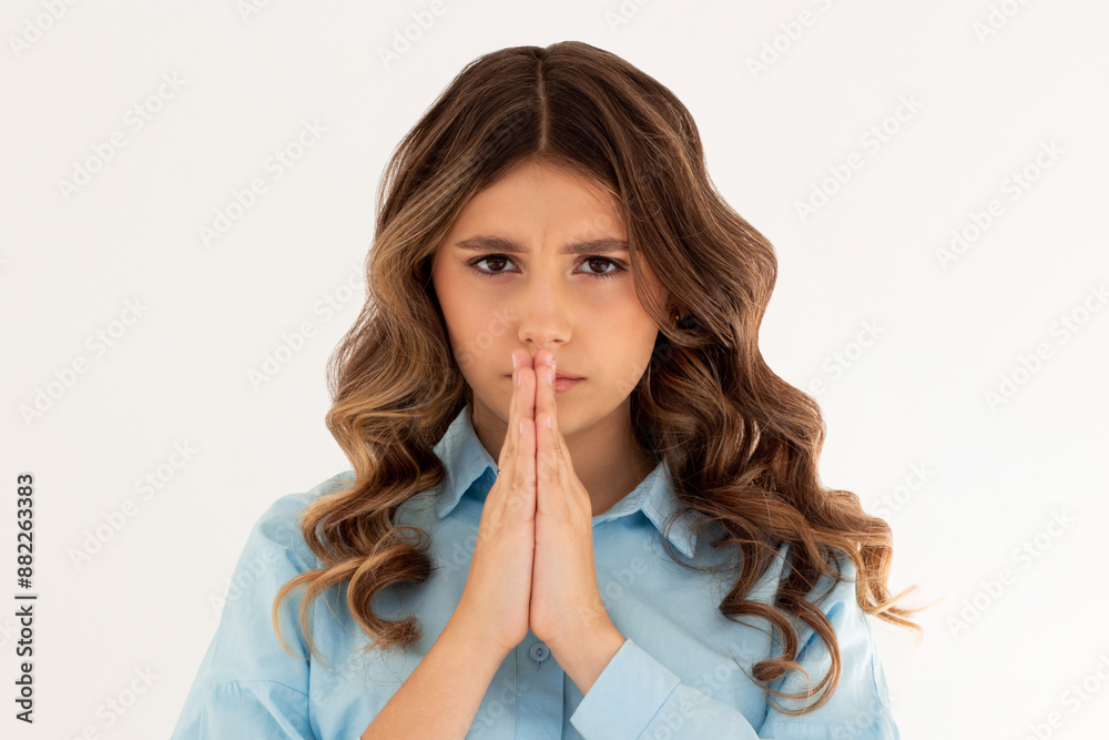 Portrait of imploring girl tilts head, purses lips, looks sadly at camera, makes praying gesture, asks for help and support, begs boyfriend for apologize, wears blue shirt, isolated on white wall
