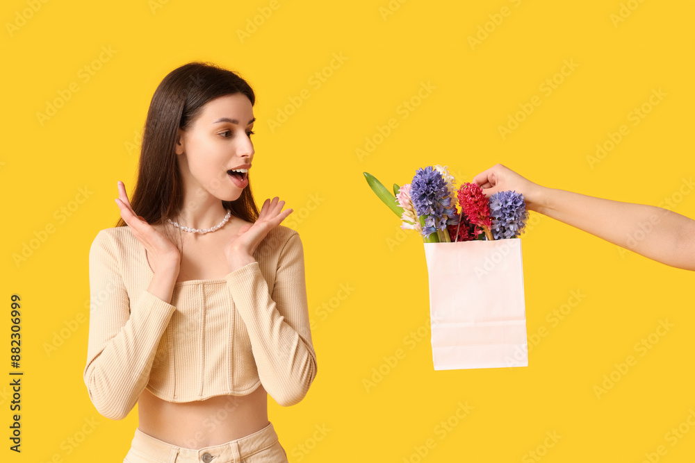 Surprised young woman and hand holding paper bag with hyacinth flowers on yellow background Surprised young woman and hand holding paper bag with hyacinth flowers on yellow background