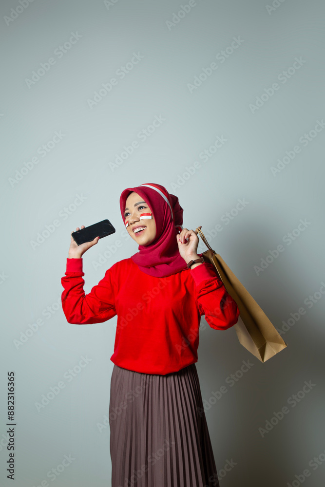 Successful Indonesian woman in hijab in red t-shirt posing candidly and carrying shopping paper bag, teenager's face with flag sticker stuck to her cheek, advertising mobile phone woman