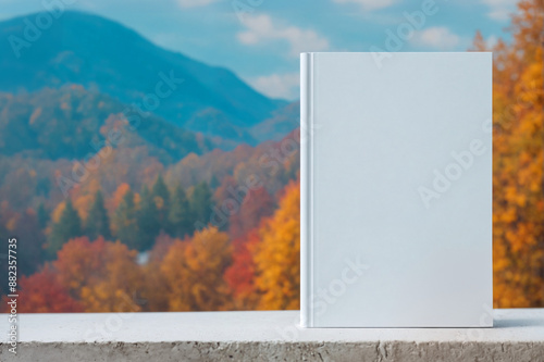 Blank white book cover mockup standing on blue wooden surface against golden, yellow autumn, fall, mountains background, landscape, outdoors. Front view of magazine template with copy space