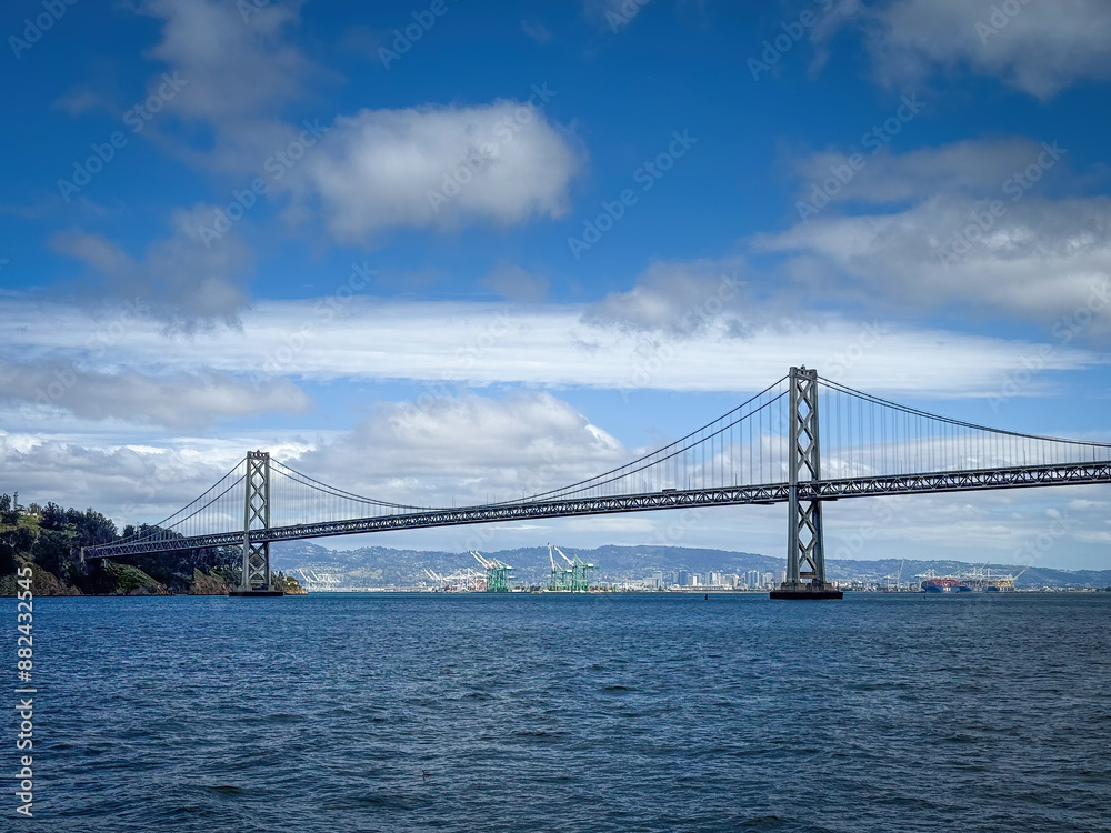 Scenic view of San Francisco-Oakland Bay Bridge and Oakland with container terminal in the background, California, USA seen from the water against blue sky with clouds Scenic view of San Francisco-Oakland Bay Bridge and Oakland with container terminal in the background, California, USA seen from the water against blue sky with clouds