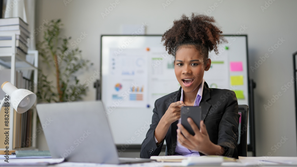 Young manager looks shocked reading a message on her phone while working in a modern office. She is african american, surrounded by charts Young manager looks shocked reading a message on her phone while working in a modern office. She is african american, surrounded by charts