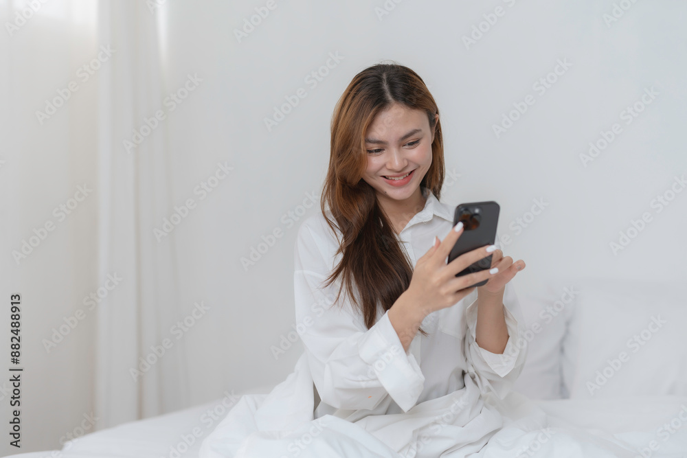 Young woman happily using her smartphone in bed, looking relaxed and content in her cozy bedroom Young woman happily using her smartphone in bed, looking relaxed and content in her cozy bedroom