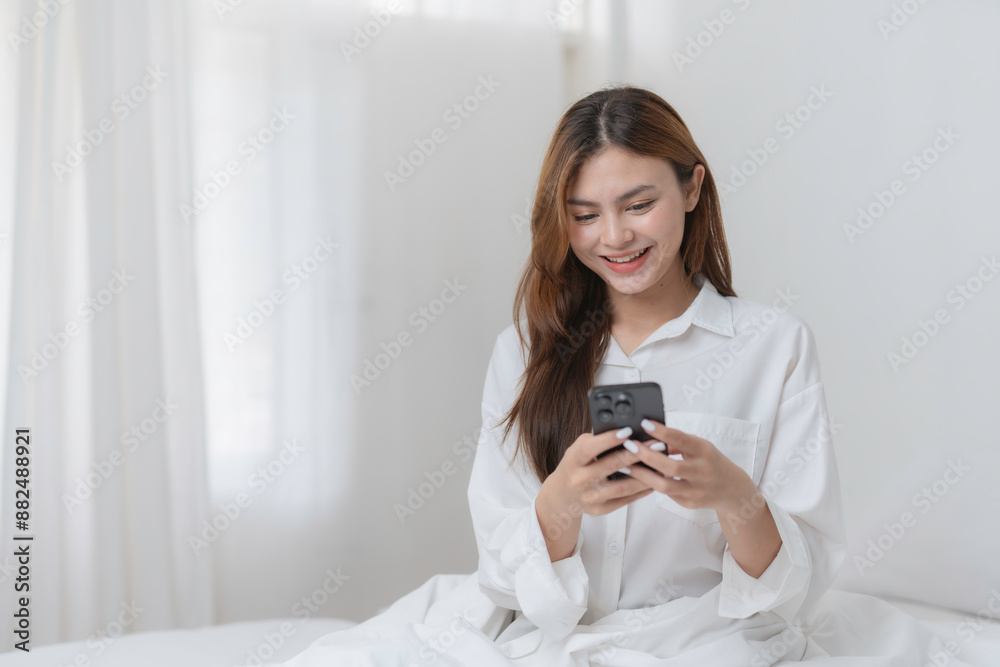 Young woman is smiling while browsing social media on her phone. She is wearing a white shirt and is sitting on a bed with white sheets Young woman is smiling while browsing social media on her phone. She is wearing a white shirt and is sitting on a bed with white sheets