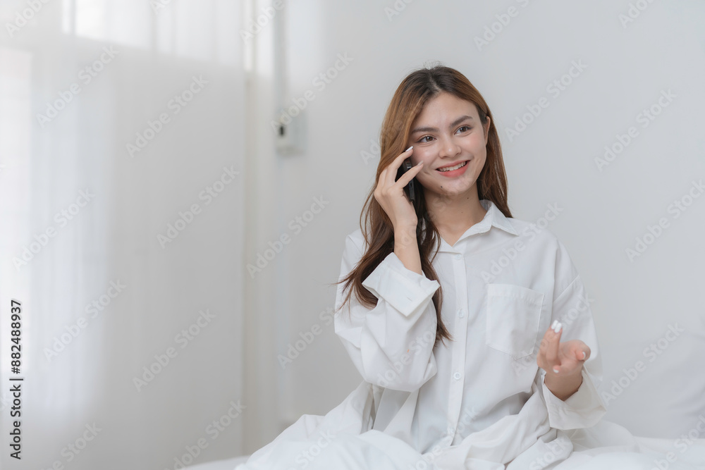 Young woman wearing a white shirt is making a phone call and smiling while sitting on a bed in a bedroom with a minimalist design Young woman wearing a white shirt is making a phone call and smiling while sitting on a bed in a bedroom with a minimalist design