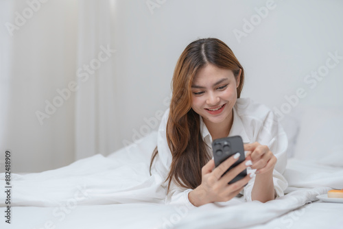 Young woman is enjoying her morning routine, lying comfortably in her bed with white sheets and using her smartphone with a smile